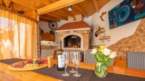 Une table en bois sur laquelle se trouve une planche de charcuterie, deux verres à vin et une bouteille de vin. On aperçoit une cheminée en briques et une horloge à l’arrière-plan.