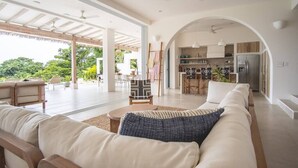 A spacious living area with a large sofa, a coffee table, and a dining area with chairs and a table. There is a kitchen visible through an archway. - Casa Niri, Luxury Villa (Guanacaste Province)