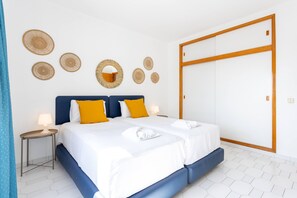 Cama doble con ropa de cama blanca, dos almohadas amarillas, cabecera de madera, mesita de noche con lámpara y un armario grande.