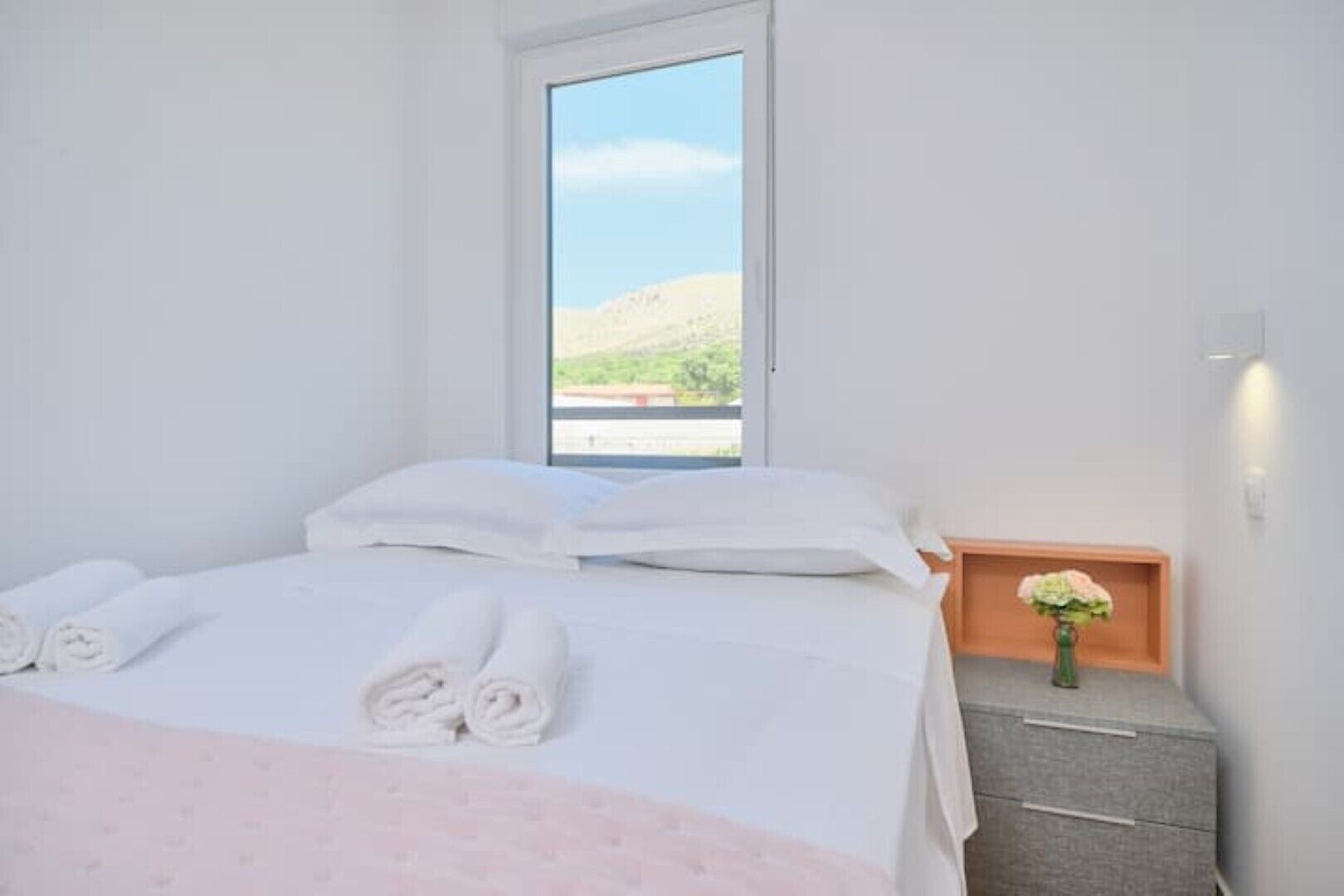 A neatly made bed with white linens and rolled towels, a small bedside table with a vase of flowers, and a window with a view of a green landscape.