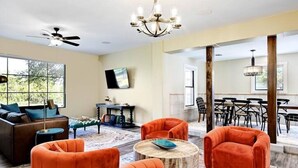 A spacious living room with a large window, a ceiling fan, and a dining area with a chandelier. - Secluded Hill Country Retreat: Bunkhouse and Spa (Austin)
