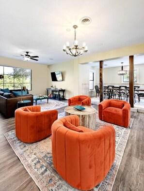A spacious living room with a large window, a ceiling fan, and a dining area with a chandelier. - Secluded Hill Country Retreat: Bunkhouse and Spa (Austin)