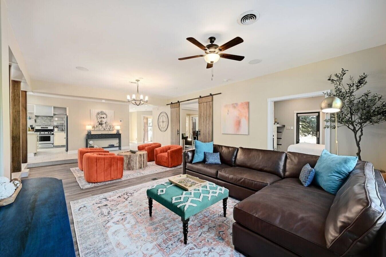 A spacious living room with a brown leather sofa, orange ottomans, and a teal ottoman. A ceiling fan and chandelier are visible.