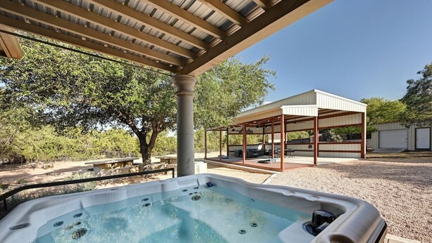 A hot tub under a pergola with a covered outdoor seating area and a building in the background. - Secluded Hill Country Retreat: Bunkhouse and Spa (Austin)