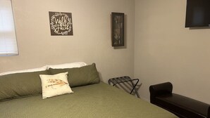 Ein Schlafzimmer mit grüner Bettdecke, einem Kopfkissen, einem kleinen Tisch und einem an der Wand befestigten Fernseher.