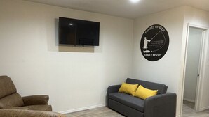 Ein Wohnzimmer mit Flachbildfernseher an der Wand, einem grauen Sofa mit gelben Kissen und einem braunen Ledersessel.