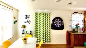 Een eetruimte met een witte tafel, gele stoelen, een groen-wit ruitpatroon gordijn, een dartbord en een muur met een inspirerende quote.