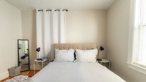 Ein Schlafzimmer mit einem großen Bett, zwei Nachttischen, einem Spiegel und einem Fenster mit Jalousien.