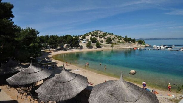 A beach with clear turquoise water, thatched umbrellas, and a small hill in the background. - MH Holiday Dream - Morning Sun (Drage)
