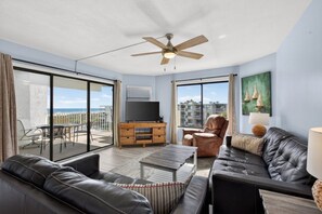 A spacious living area with a large window, a ceiling fan, a flat-screen TV, a leather sofa, a coffee table, and a dining table with chairs. - Relaxation Reef - Ocean Views, Heated Pool, Lovely Condo, Beach Access (St. Augustine)