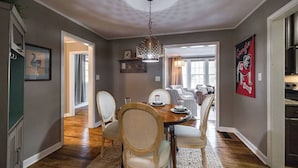 A dining area with a wooden table, chairs, and a chandelier. There is a view into another room with a couch and a window. - Five Points Charmer: Delightful 3BR Cottage in Five Points, Complete W/Fire Pit. (Athens)