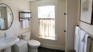A bathroom with a toilet, sink, and a window with curtains. There are towels on a shelf and a framed picture on the wall.