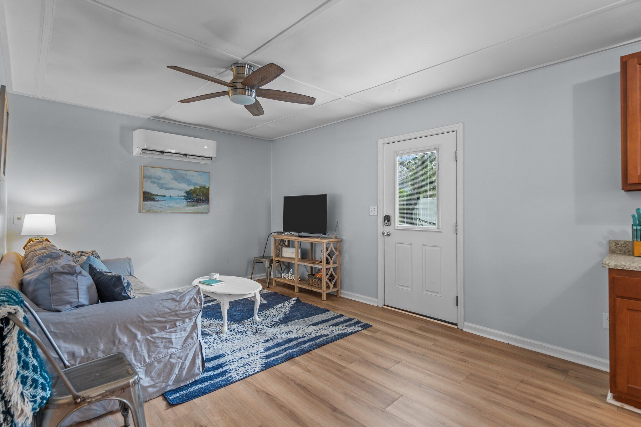 Ein Wohnzimmer mit Deckenventilator, Fernseher auf einem Holzboden, einer Couch, einem Couchtisch und einer Tür mit Fenster.