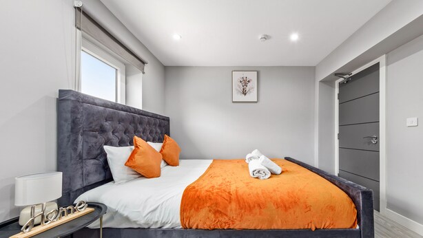 Een moderne slaapkamer met een groot bed, een grijs gestoffeerd hoofdeinde, beddengoed in wit en oranje, een nachtkastje met een lamp en een lijst met een kunstwerk aan de muur.