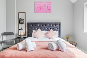 A neatly arranged bedroom with a tufted headboard, a pink bedspread, and two rolled towels on the bed.