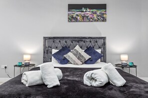 Une chambre à coucher moderne avec une tête de lit capitonnée, deux tables de chevet avec des lampes et un grand tableau au-dessus du lit.