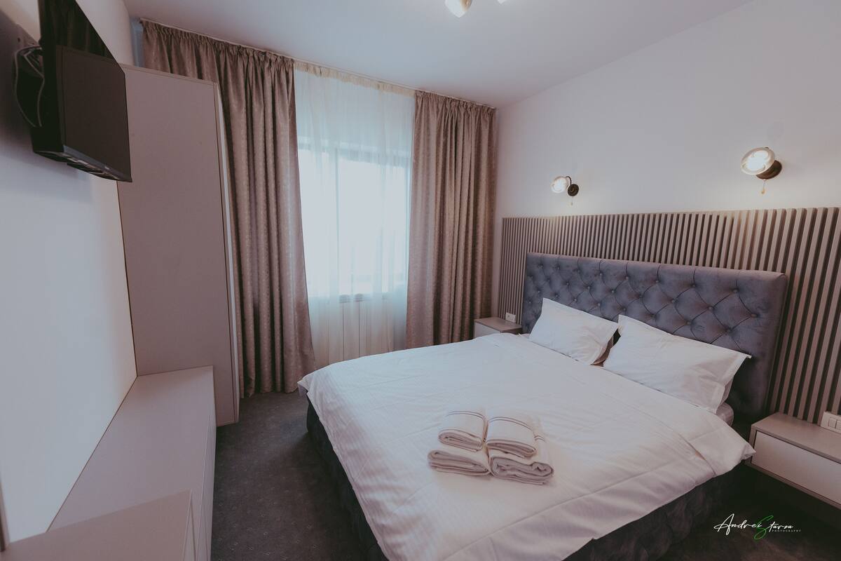 A hotel room with a large bed, a flat-screen TV mounted on the wall, a bedside table, and a window with curtains.