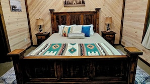 Ein Schlafzimmer mit Holzbalkendecke, einem Bett mit gemusterten Betttextilien, zwei Nachttischen mit Lampen und einem Wandbild.