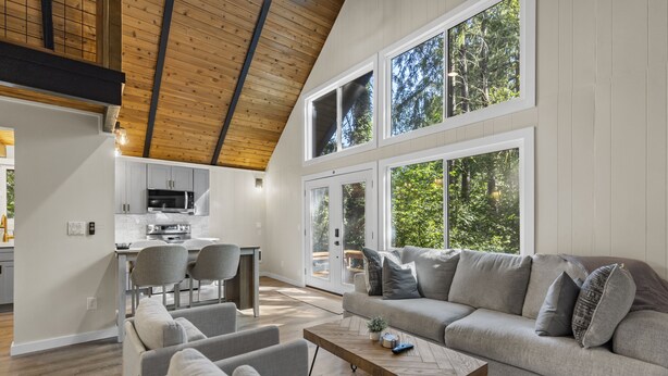 Un interno moderno con soffitto a loft, una zona giorno con divano e poltrone, una cucina con forno a microonde e ampie finestre che si affacciano su alberi.