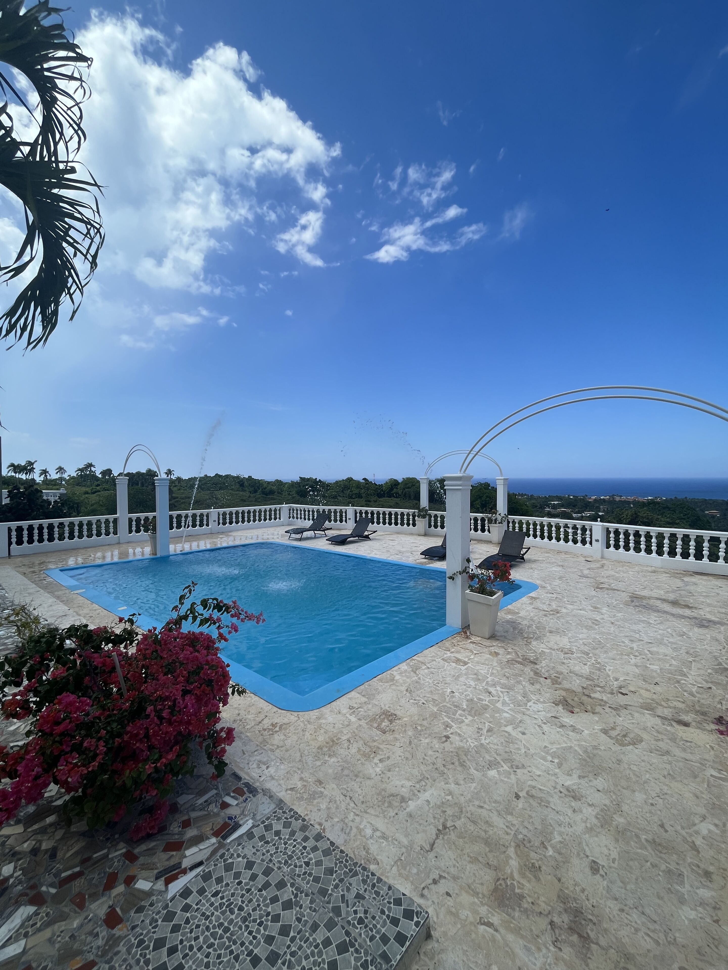 A pool area with a clear view of the ocean, surrounded by a white balustrade and lounge chairs.