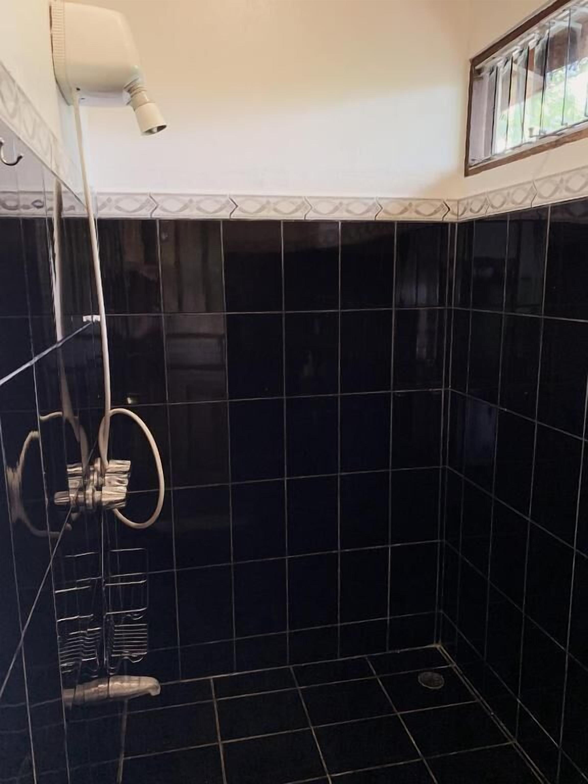 A small, dark-tiled bathroom with a handheld showerhead, a towel rack, and a window with bars.