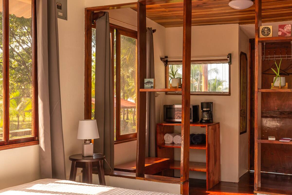 A bedroom with a large bed, wooden beams, a desk with a lamp, and a view of the outdoors.