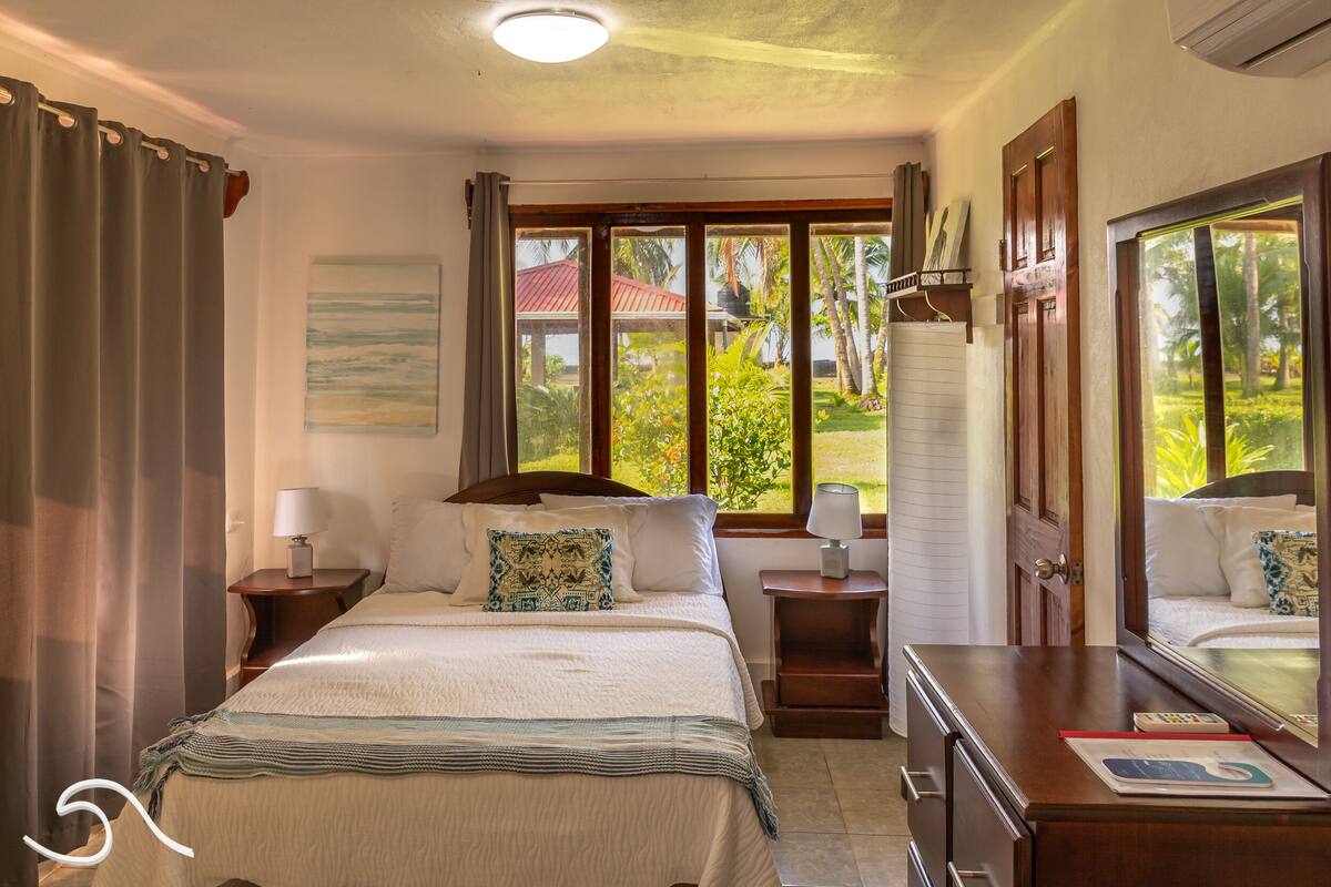 A hotel room with a bed, two bedside tables, a dresser, and a large window offering a view of greenery and a building with a red roof.