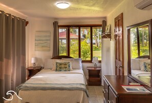 A hotel room with a bed, two bedside tables, a dresser, and a large window offering a view of greenery and a building with a red roof.
