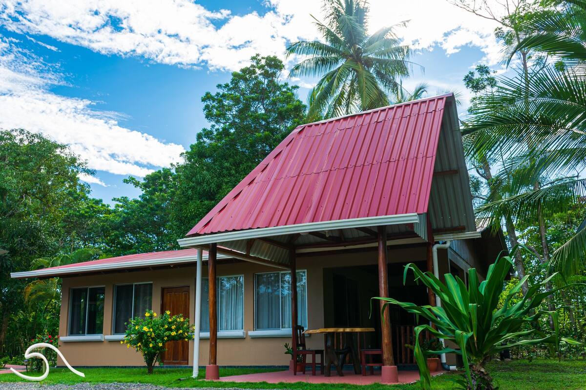 A two-story house with a red roof, a porch with wooden chairs, and a small garden area.