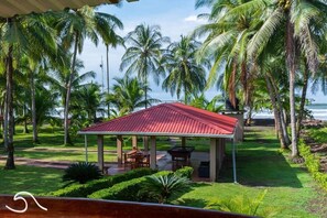 A covered outdoor seating area with a red roof, surrounded by palm trees and a view of the ocean.