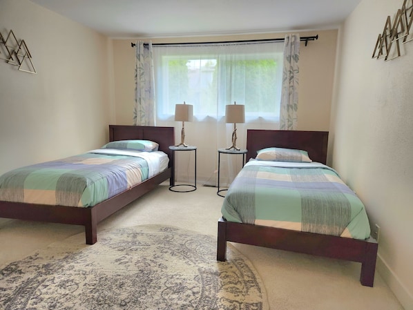 Un dormitorio con dos camas individuales, una mesita redonda con lámpara, una ventana con cortinas traslúcidas y una alfombra estampada.