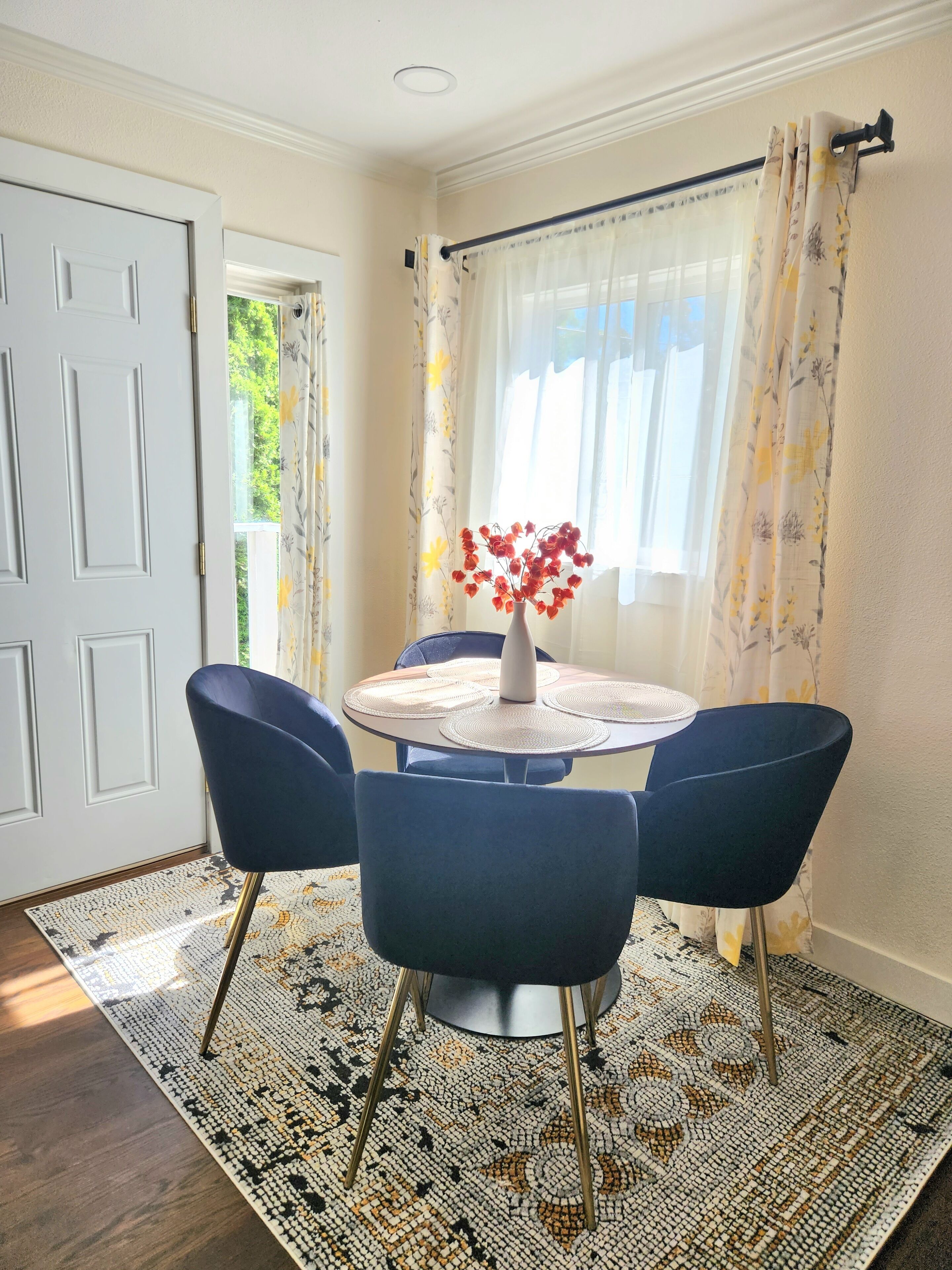 Un coin repas avec une table ronde, quatre chaises bleues, un vase avec des fleurs rouges et un tapis à motifs.