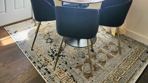 Un coin repas avec une table ronde, quatre chaises bleues, un vase avec des fleurs rouges et un tapis à motifs.