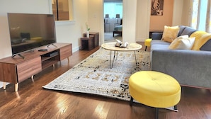 Un salon moderne doté d’un téléviseur à écran plat, de meubles en bois, d’un tapis à motifs et d’un canapé agrémenté de coussins jaunes.