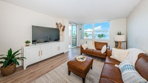 Un salon équipé d’un téléviseur, d’un canapé, d’un pouf et offrant une vue sur une étendue d’eau par une fenêtre.