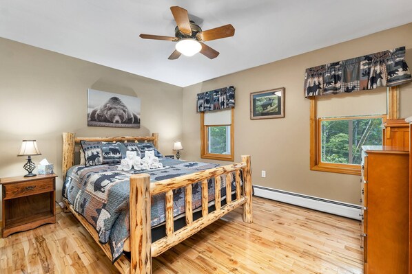 A bedroom with a wooden bed, a ceiling fan, a window with a view of trees, and a bear-themed wall hanging.