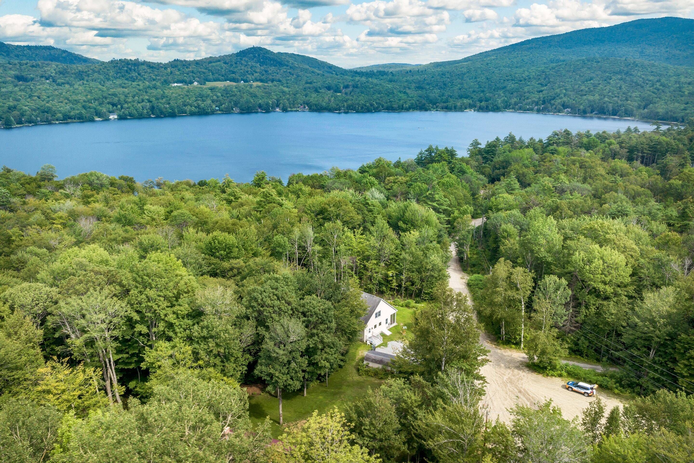 An aerial view of a lake surrounded by dense forest and hills. A white house is visible in the foreground with a dirt path leading to it.