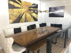 A dining area with a long wooden table, white upholstered chairs, and black cushions. There are two wall-mounted paintings above the table.