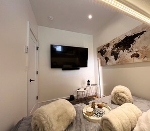 Una habitación moderna con un televisor de pantalla plana montado en la pared, una mesita redonda con una bandeja y dos otomanas blancas y mullidas.