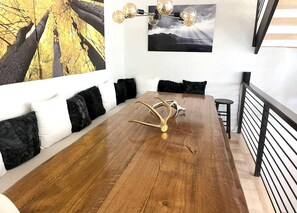 Un comedor con una larga mesa de madera, cojines blancos y negros, y una lámpara de araña moderna.