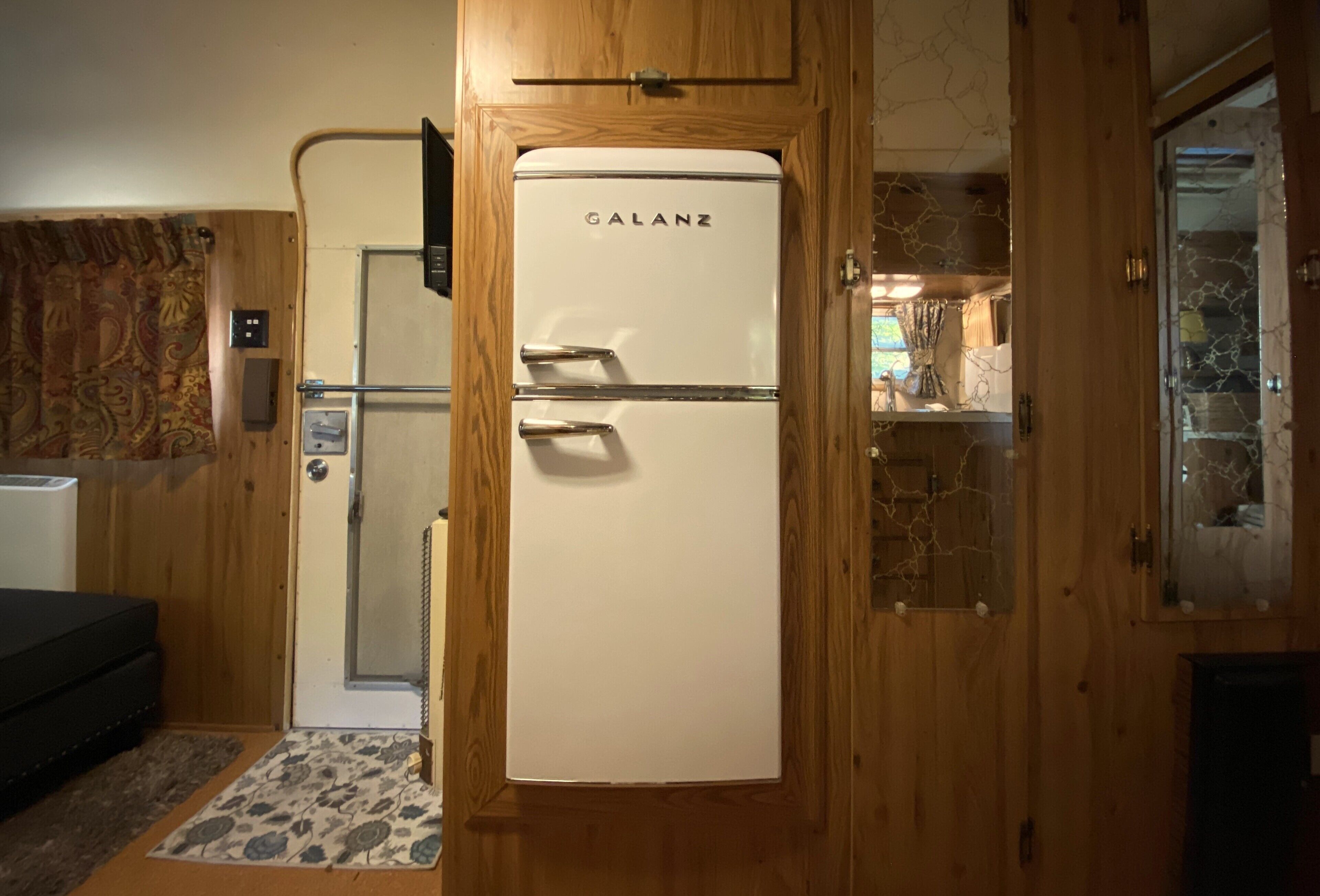 A compact room with a wooden door, a white Galanz refrigerator, a small bed, and a patterned rug.