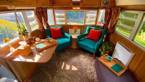 En hyggelig autocamper-indretning med to teal farvede lænestole, et lille bord med en vase blomster, og en lilla sofa.