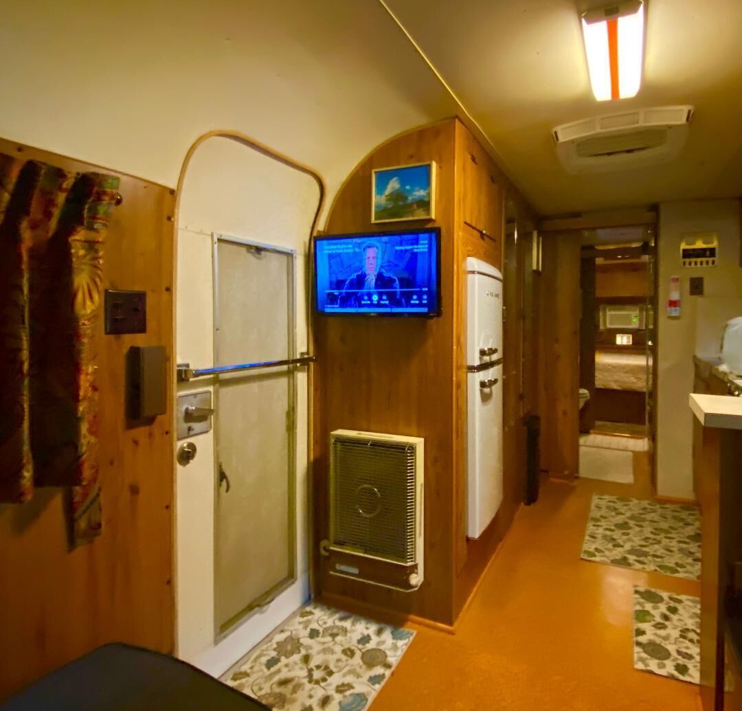 Ein schmaler Flur mit an der Wand montiertem Fernseher, einem Kühlschrank und einer Tür, vor der ein kleiner Teppich liegt.
