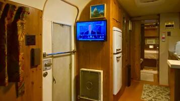 Ein schmaler Flur mit an der Wand montiertem Fernseher, einem Kühlschrank und einer Tür, vor der ein kleiner Teppich liegt.