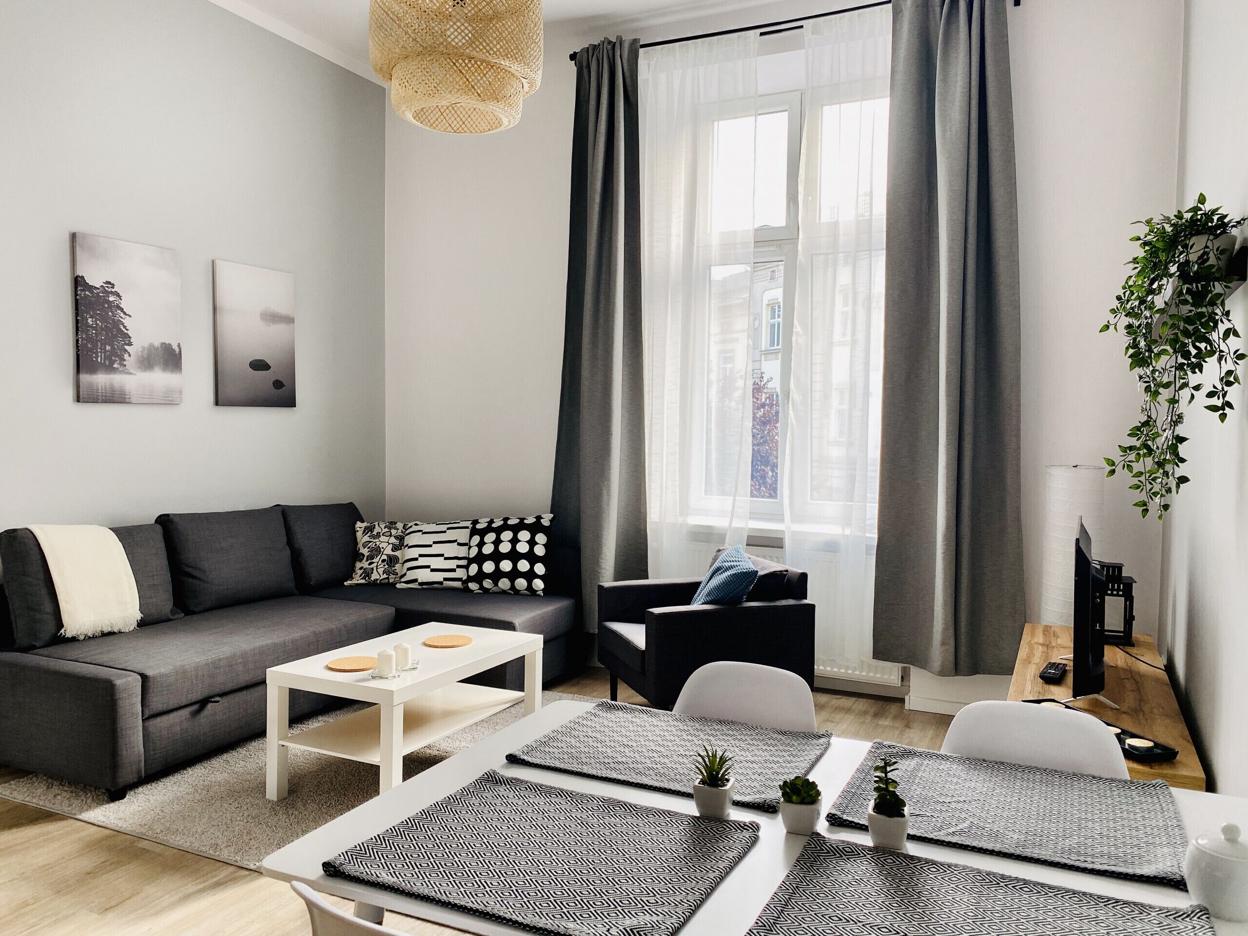 Un salon moderne avec un canapé gris, une table basse blanche et un fauteuil noir. Il y a deux fenêtres avec des rideaux gris, un luminaire suspendu en osier et une petite plante en pot sur la droite.