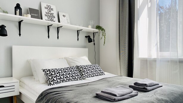 Ein modernes Schlafzimmer mit einem weißen Bett, grauer Bettwäsche und einer grauen Decke. Es gibt zwei Nachttische, einer mit einer Lampe und der andere mit einer kleinen Pflanze. Ein Wandregal beherbergt dekorative Gegenstände und ein gerahmtes Bild.