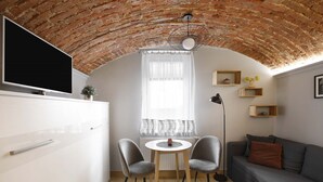 A modern living room with a brick ceiling, a flat-screen TV mounted on the wall, a round dining table with two chairs, a grey sofa, and a floor lamp.