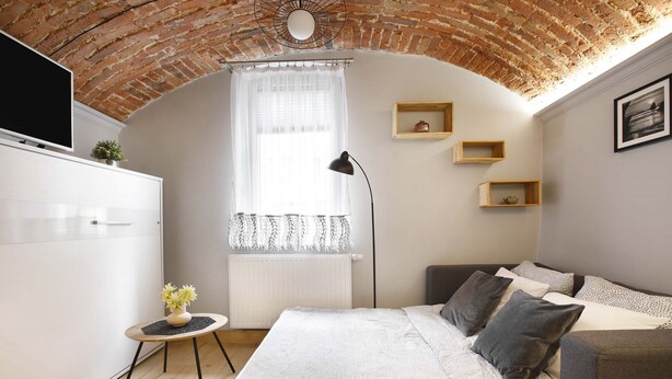 A cozy bedroom with a brick ceiling, a large bed with grey and white bedding, a small side table with a plant, and a window with sheer curtains.