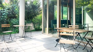 Een modern buitenterras met houten tafels en stoelen, en grote glazen deuren die uitkomen op een groene tuin.