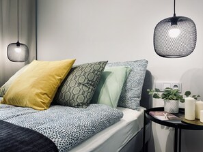 Cama bem arrumada com roupa de cama estampada, mesa de cabeceira com planta em vaso e duas luminárias pendentes.